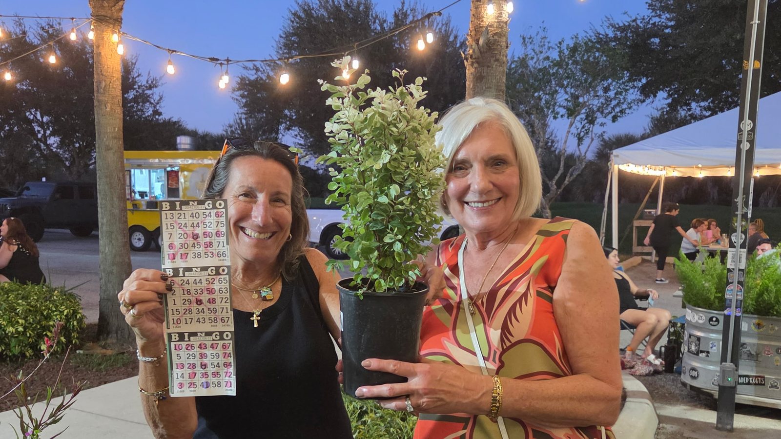 Two smiling winners at Plant Bingo under string lights, one holding her bingo card and the other her prize plant
